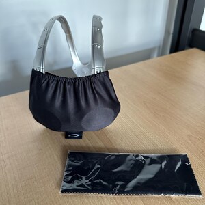 May include: Silver-framed sunglasses with a black fabric pouch and a black cleaning cloth. The sunglasses have a distinctive curved frame and a black lens cover. The cleaning cloth is in a clear plastic wrapper.