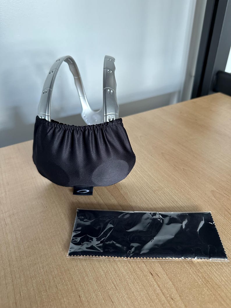 May include: Silver-framed sunglasses with a black fabric pouch and a black cleaning cloth. The sunglasses have a distinctive curved frame and a black lens cover. The cleaning cloth is in a clear plastic wrapper.