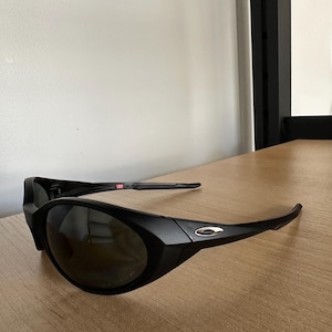 Puede incluir: Gafas de sol negras con un diseño curvo y lentes oscuras. Las gafas tienen un acabado mate y un pequeño logotipo plateado en el lateral. La montura tiene un sutil detalle rojo, blanco y azul.