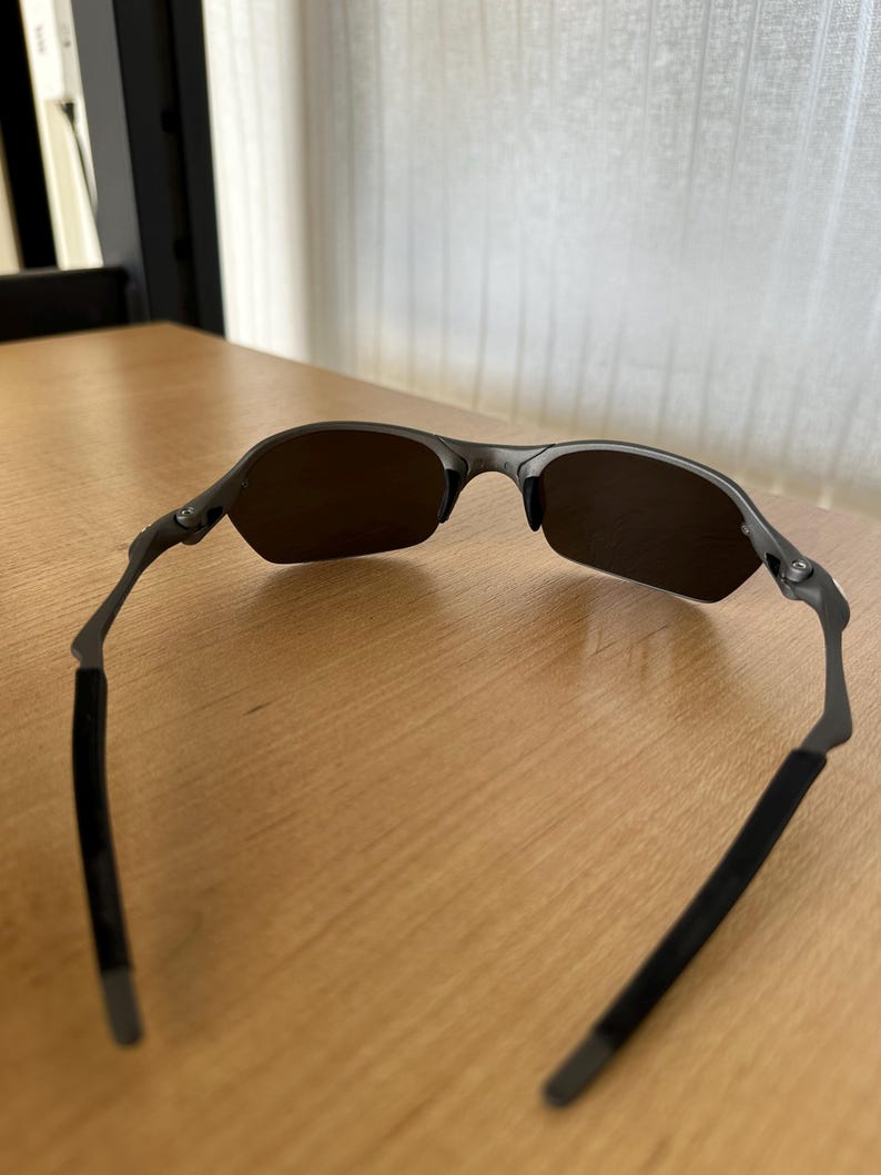 Pode incluir: &Oacute;culos de sol cinza escuro com um design semi-sem aro. As lentes s&atilde;o de cor marrom escuro e as arma&ccedil;&otilde;es s&atilde;o cinza fosco. As hastes t&ecirc;m um revestimento de borracha preta para conforto.