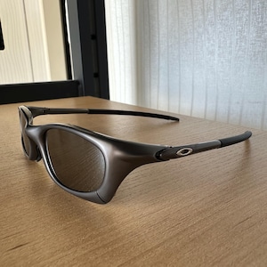 May include: Dark grey sunglasses with a curved frame and tinted lenses. The sunglasses are on a light brown wooden surface. The temples are thin and extend back to the ear pieces.