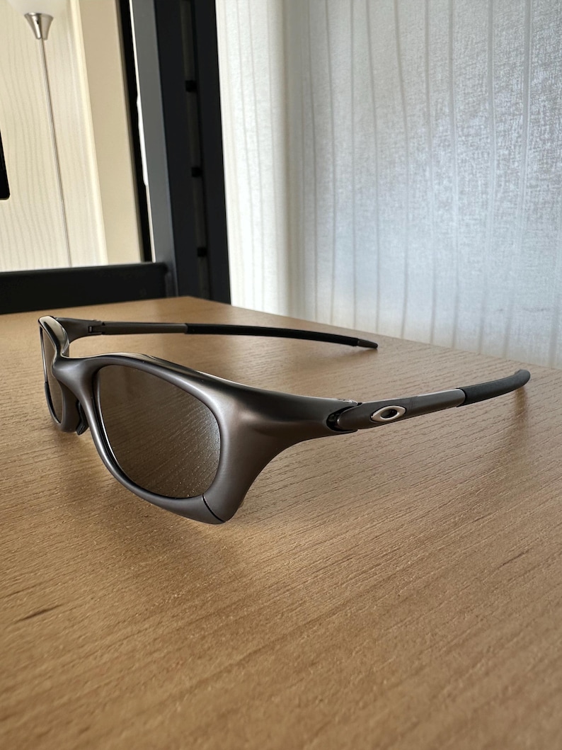 May include: Dark grey sunglasses with a curved frame and tinted lenses. The sunglasses are on a light brown wooden surface. The temples are thin and extend back to the ear pieces.
