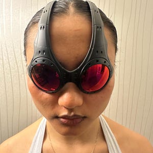 May include: Black sport sunglasses with red lenses. The sunglasses have a unique design with a thick frame that extends over the forehead. The lenses are a vibrant red color, and the frame has a matte finish.