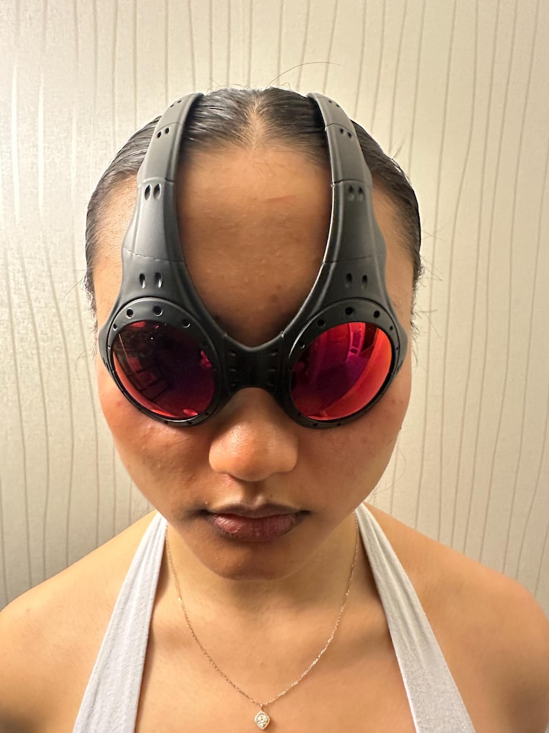 May include: Black sport sunglasses with red lenses. The sunglasses have a unique design with a thick frame that extends over the forehead. The lenses are a vibrant red color, and the frame has a matte finish.