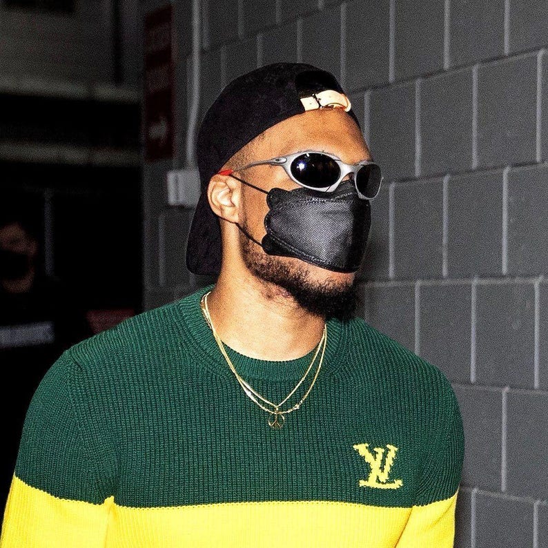 May include: A person wearing a black baseball cap, silver-framed sunglasses, and a black face mask. They are wearing a green and yellow sweater with a gold logo and a gold necklace.