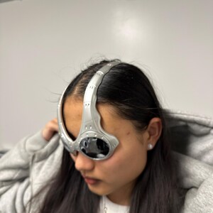 May include: Silver futuristic eyewear with dark lenses. The glasses have a curved design that wraps around the forehead and temples. The eyewear is made of a metallic material and has a sleek, modern aesthetic.