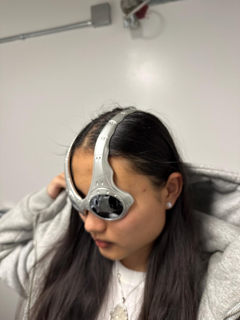 May include: Silver futuristic eyewear with dark lenses. The glasses have a curved design that wraps around the forehead and temples. The eyewear is made of a metallic material and has a sleek, modern aesthetic.