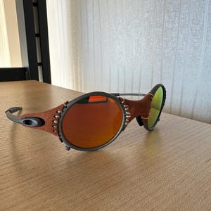 May include: Oval-shaped sunglasses with a brown leather and gray frame. The lenses are a gradient orange color. The temples are gray and the bridge is a thin metal. The sunglasses are on a light brown wooden surface.
