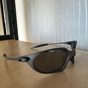 May include: Gray sunglasses with dark lenses. The sunglasses have a sleek, modern design with a curved frame and a distinctive shape. The arms are thin and extend to the temples. The lenses are oval-shaped.