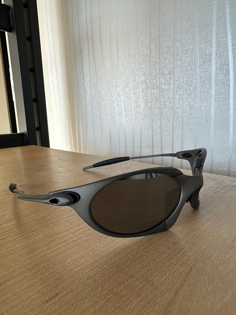 May include: Gray sunglasses with dark lenses. The sunglasses have a sleek, modern design with a curved frame and a distinctive shape. The arms are thin and extend to the temples. The lenses are oval-shaped.