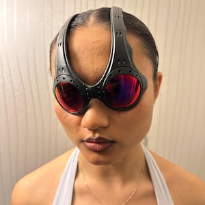 May include: Black sport sunglasses with red-tinted lenses. The sunglasses have a unique, wraparound design with a thick frame and a bridge that extends over the top of the head. The lenses are round and the frame has a matte finish.