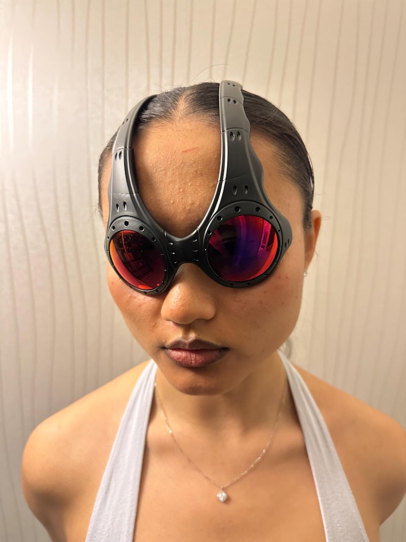 May include: Black sport sunglasses with red-tinted lenses. The sunglasses have a unique, wraparound design with a thick frame and a bridge that extends over the top of the head. The lenses are round and the frame has a matte finish.