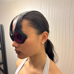 May include: Black and red-tinted goggles with a thick, curved black frame. The goggles have a wraparound design and are worn on the head. The lenses are a deep red color.
