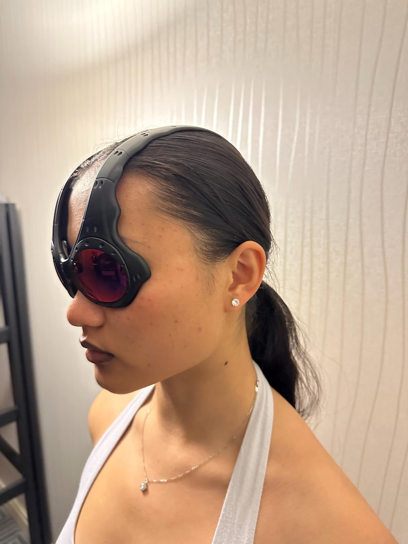 May include: Black and red-tinted goggles with a thick, curved black frame. The goggles have a wraparound design and are worn on the head. The lenses are a deep red color.