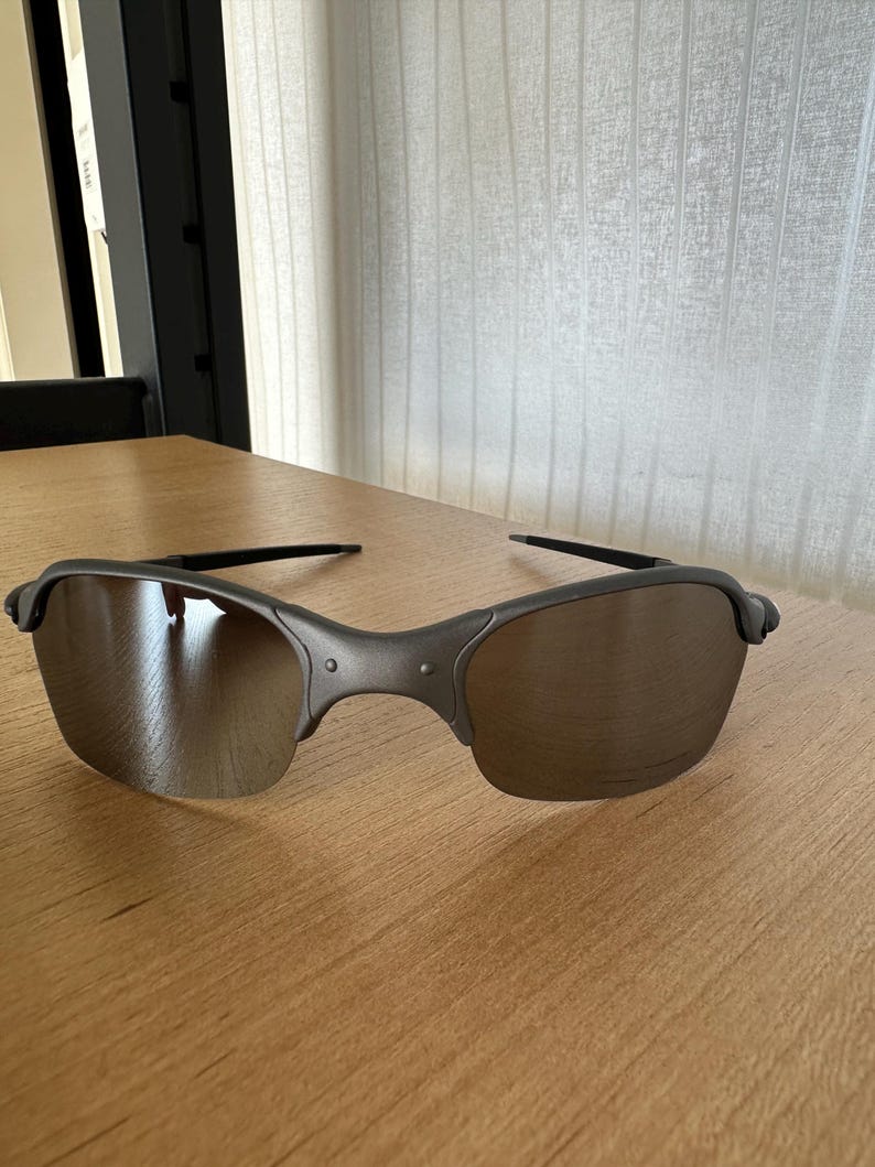 Pode incluir: Um par de &oacute;culos de sol com arma&ccedil;&atilde;o cinza fosco e lentes tingidas de marrom. Os &oacute;culos de sol est&atilde;o sobre uma superf&iacute;cie de madeira marrom clara. As lentes t&ecirc;m uma ligeira curvatura.