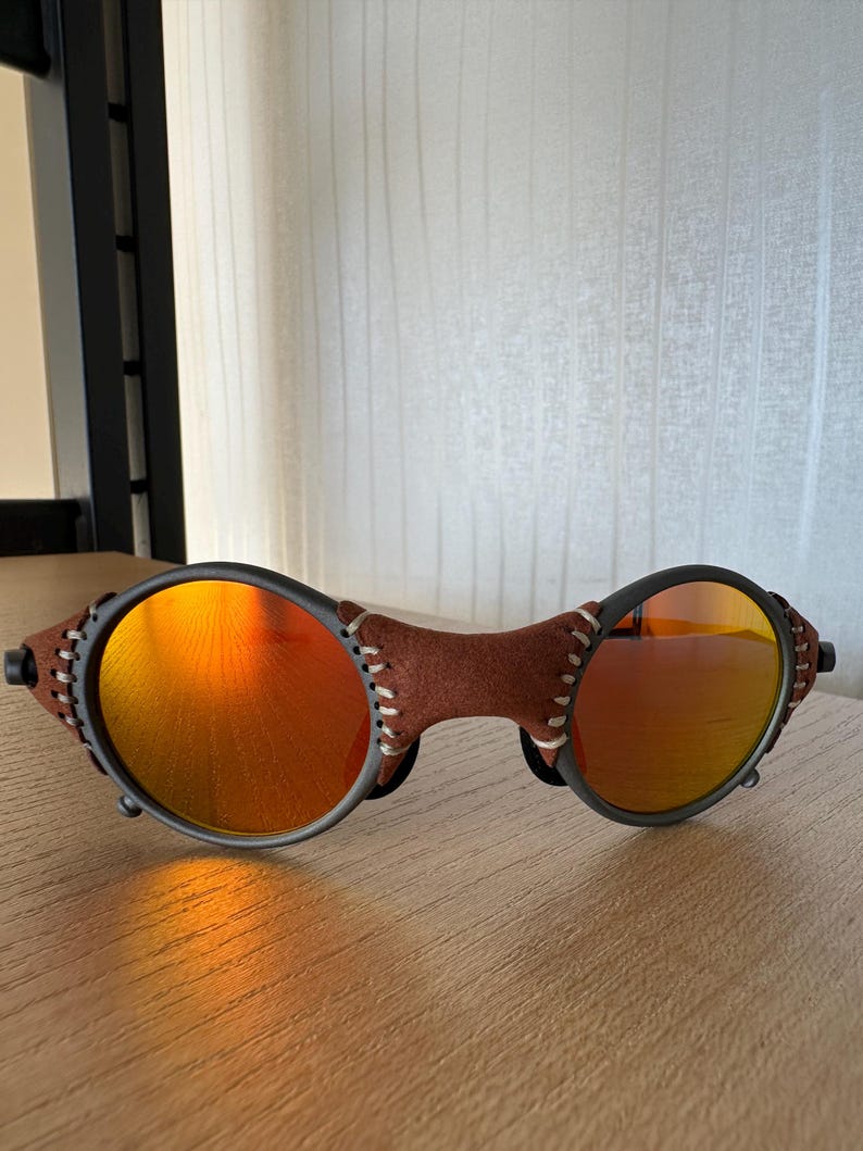 May include: Sunglasses with orange lenses and a brown leather bridge. The frames are black with white stitching on the leather. The lenses reflect a bright orange color.