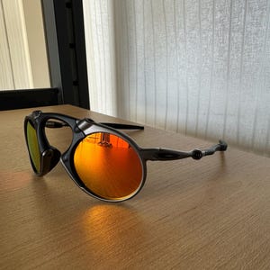 Pu&ograve; includere: Occhiali da sole neri e grigi con lenti a specchio arancioni. Gli occhiali hanno una montatura curva e un design moderno. Le lenti riflettono l'ambiente, creando una vibrante tonalit&agrave; arancione.