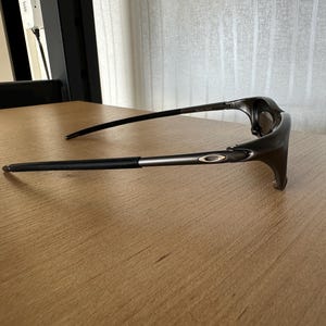 May include: Dark grey sunglasses with black arms and a silver accent. The glasses have a sleek, modern design and the Oakley logo is visible on the arm.