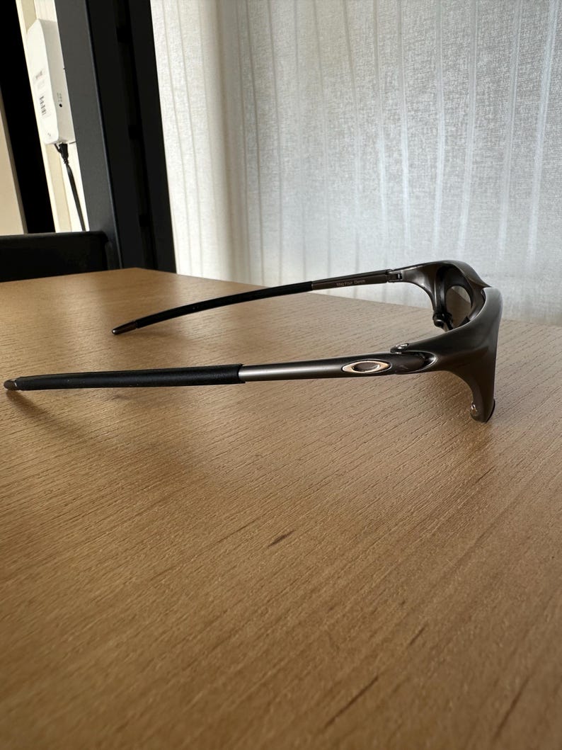 May include: Dark grey sunglasses with black arms and a silver accent. The glasses have a sleek, modern design and the Oakley logo is visible on the arm.