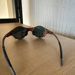 May include: Sunglasses with a brown leather and metal frame. The lenses are round and dark, with a green tint. The temples are dark gray with black rubber ear pieces. The sunglasses are resting on a light brown wooden surface.