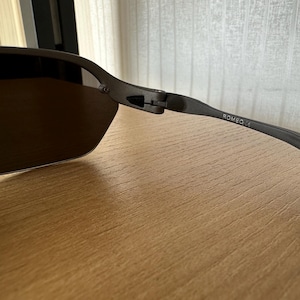 Pode incluir: &Oacute;culos de sol cinza escuro com lentes marrons. A arma&ccedil;&atilde;o tem um acabamento fosco e na haste dos &oacute;culos est&aacute; a palavra "ROMEO". Os &oacute;culos de sol est&atilde;o sobre uma superf&iacute;cie de madeira.