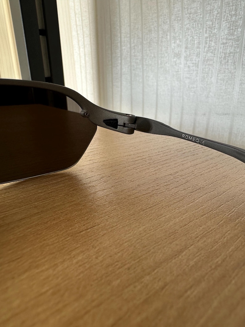 Pode incluir: &Oacute;culos de sol cinza escuro com lentes marrons. A arma&ccedil;&atilde;o tem um acabamento fosco e na haste dos &oacute;culos est&aacute; a palavra "ROMEO". Os &oacute;culos de sol est&atilde;o sobre uma superf&iacute;cie de madeira.