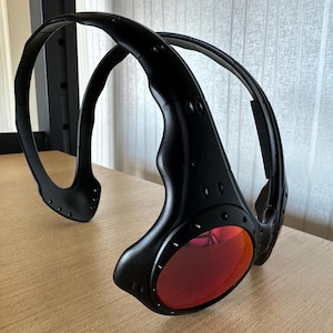 May include: Black sunglasses with red lenses. The sunglasses have a unique wraparound design with a thick, curved frame. The lenses are oval-shaped and the frame has a matte finish. The sunglasses are resting on a wooden surface.