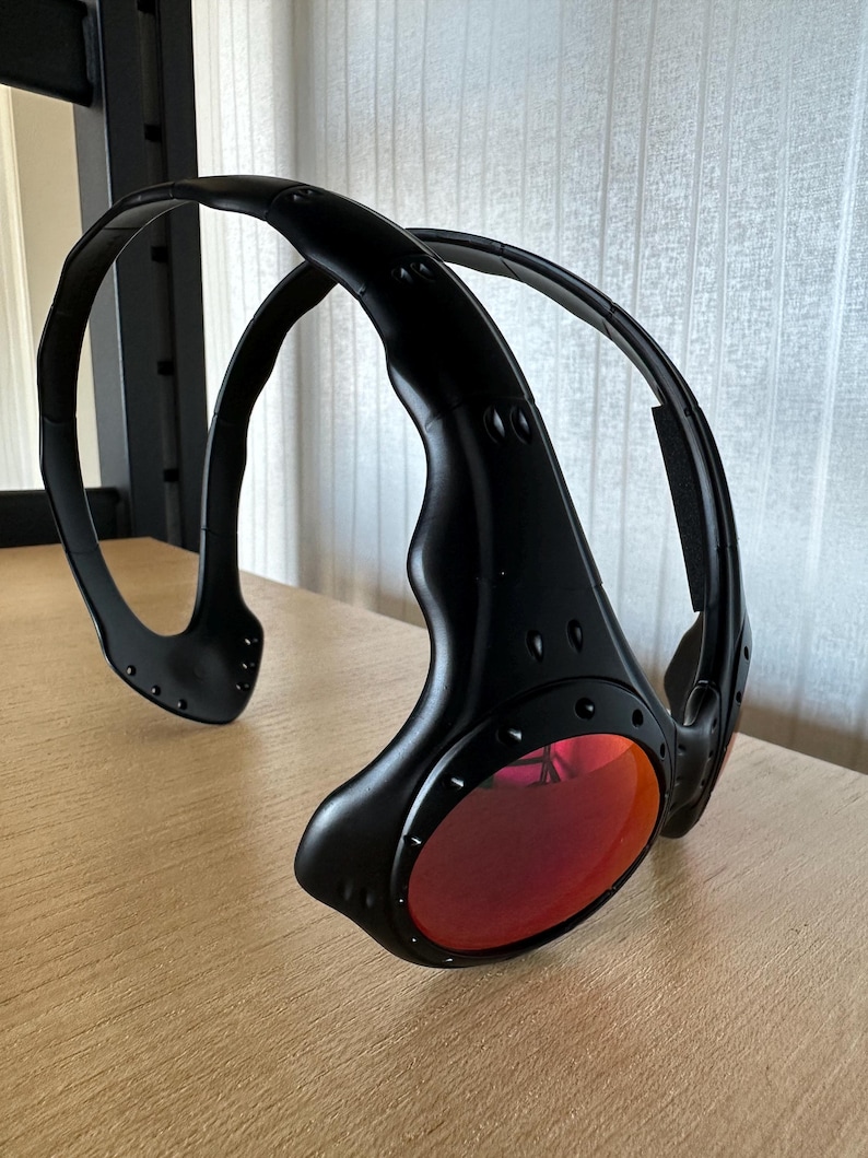 May include: Black sunglasses with red lenses. The sunglasses have a unique wraparound design with a thick, curved frame. The lenses are oval-shaped and the frame has a matte finish. The sunglasses are resting on a wooden surface.