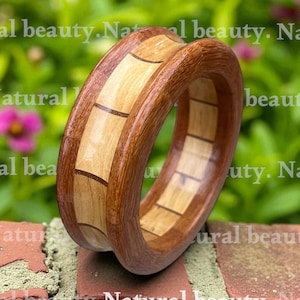 May include: A wooden ring with a natural finish. The ring features alternating light and dark wood segments, creating a geometric pattern. The ring is displayed on a brick surface with a green background.