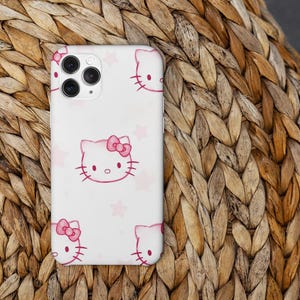 May include: A white phone case with a repeating pattern of pink Hello Kitty faces and pink stars. The case has a camera cutout and is set against a woven, light brown background.