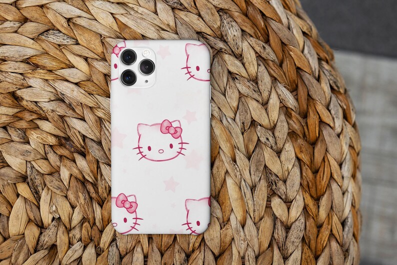 May include: A white phone case with a repeating pattern of pink Hello Kitty faces and pink stars. The case has a camera cutout and is set against a woven, light brown background.