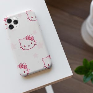 May include: A white phone case with a pink Hello Kitty design, including a bow and whiskers, and pink stars. The case is on a white surface, with a blurred background including a statue and greenery.