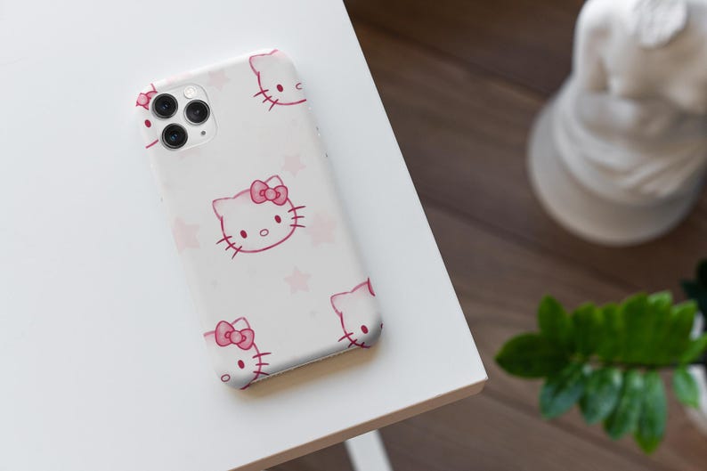 May include: A white phone case with a pink Hello Kitty design, including a bow and whiskers, and pink stars. The case is on a white surface, with a blurred background including a statue and greenery.