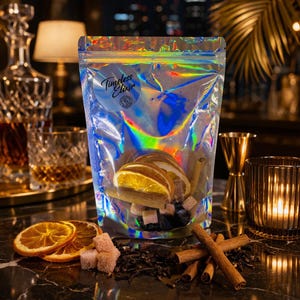 Op de afbeelding: Een holografische zak met het label "Timeless Elixir" gevuld met gedroogde sinaasappelschijfjes, suikerklontjes en kaneelstokjes. De zak staat op een marmeren oppervlak met extra sinaasappelschijfjes, suikerklontjes en kaneelstokjes.