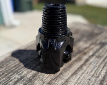 May include: A black metal drill bit with a threaded top and a multi-faceted cutting head. The bit is designed for industrial use and sits on a weathered wooden surface. The image is taken in natural light.