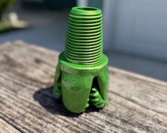May include: A metallic green drill bit, featuring a threaded top and a unique, claw-like base. The tool is designed for drilling and is made of a durable material. The image is taken outdoors on a wooden surface.