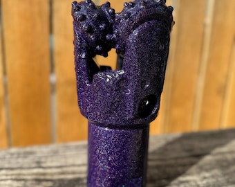 May include: A close-up of a purple drill bit with a glittery finish. The drill bit has a cylindrical base and a head with multiple rounded protrusions. The background is a wooden fence.