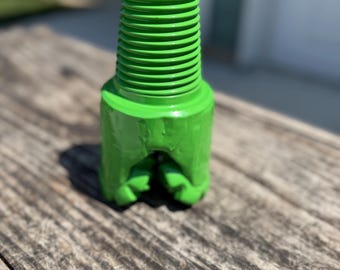 May include: Bright green drill bit with a threaded top and a toothed bottom. The tool is made of metal and sits on a weathered wooden surface. The drill bit is designed for industrial use.