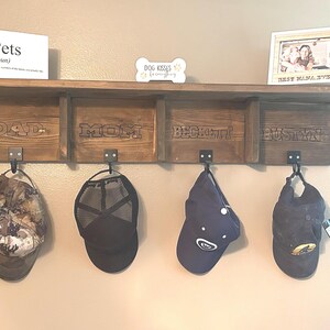 May include: A wooden wall shelf with four compartments, each labeled with a name. Below, four hats hang from black metal hooks. Above, a sign reads "Pets" and a small framed photo.