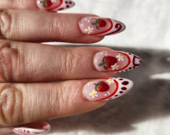 May include: Close-up of a hand with almond-shaped nails painted with a white base and red strawberry designs. The nails have a white and red swirl pattern with white dots and small white flowers.