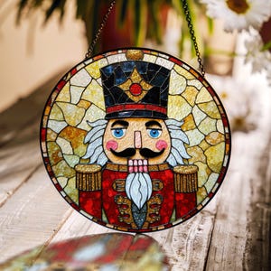 May include: A stained glass nutcracker ornament with a red, white, and blue design. The nutcracker is wearing a black hat with gold accents and has a white beard.