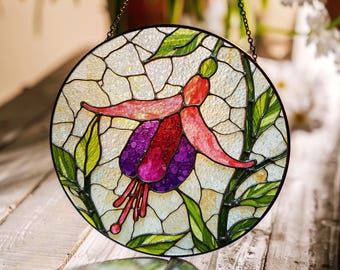 May include: A round stained glass window featuring a fuchsia flower design. The flower has red, purple, and pink petals and is surrounded by green leaves and stems. The glass has a textured, crackled appearance, and the window is framed in black with a hanging chain.