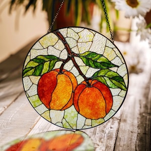 May include: Circular stained glass panel depicting two peaches on a branch with green leaves. The peaches are orange and red, set against a background of textured, light-coloured glass. A decorative piece with a dark metal chain for hanging.