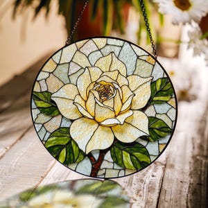 Puede incluir: Vidriera redonda que representa una gran flor amarilla con hojas verdes y un tallo marrón. La flor es el punto central, rodeada de un mosaico de vidrio claro. Una cadena negra está adjunta para colgarla.
