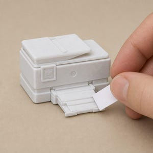 May include: A miniature white 3D printed printer model featuring a flat top lid, a front control panel with a small square button, and an extended paper tray. A small, plain white rectangular piece of paper is positioned at the printer output slot.