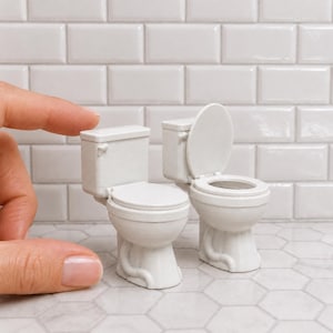 May include: Two miniature white toilets made of plastic. One toilet has the lid closed, while the other toilet has the lid open, revealing the bowl. These small bathroom fixtures are designed for dollhouse displays or dioramas and are white in color.