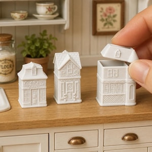 May include: Three miniature white ceramic house-shaped containers with intricate architectural details including windows, doors, and roof textures. One container has its lid removed, revealing a hollow interior space for storage or display.