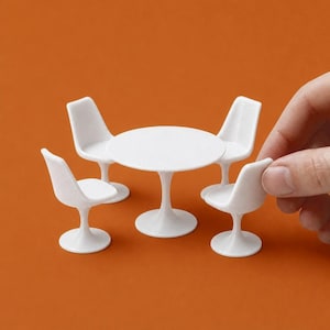 Op de afbeelding: Een witte miniatuurtafel met een rond blad en vier bijpassende stoelen. De stoelen en de tafel hebben een voet. De set staat op een oranje achtergrond. Een hand houdt een van de stoelen vast.