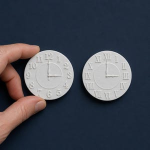 Peut inclure: Deux cadrans d'horloge circulaires blancs avec des détails en relief. L'un présente des chiffres arabes de 1 à 12, tandis que l'autre affiche des chiffres romains de I à XII. Les deux ont des aiguilles indiquant trois heures précises.
