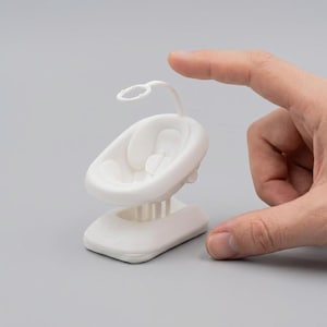 Könnte beinhalten: Ein kleiner, weißer Baby-Wippstuhl mit einem geschwungenen Sitz, einem Mobile und einem Sockel. Der Wippstuhl wird von einer Hand vor grauem Hintergrund gehalten. Der Sitz hat eine Kopfstütze und einen Sicherheitsgurt.